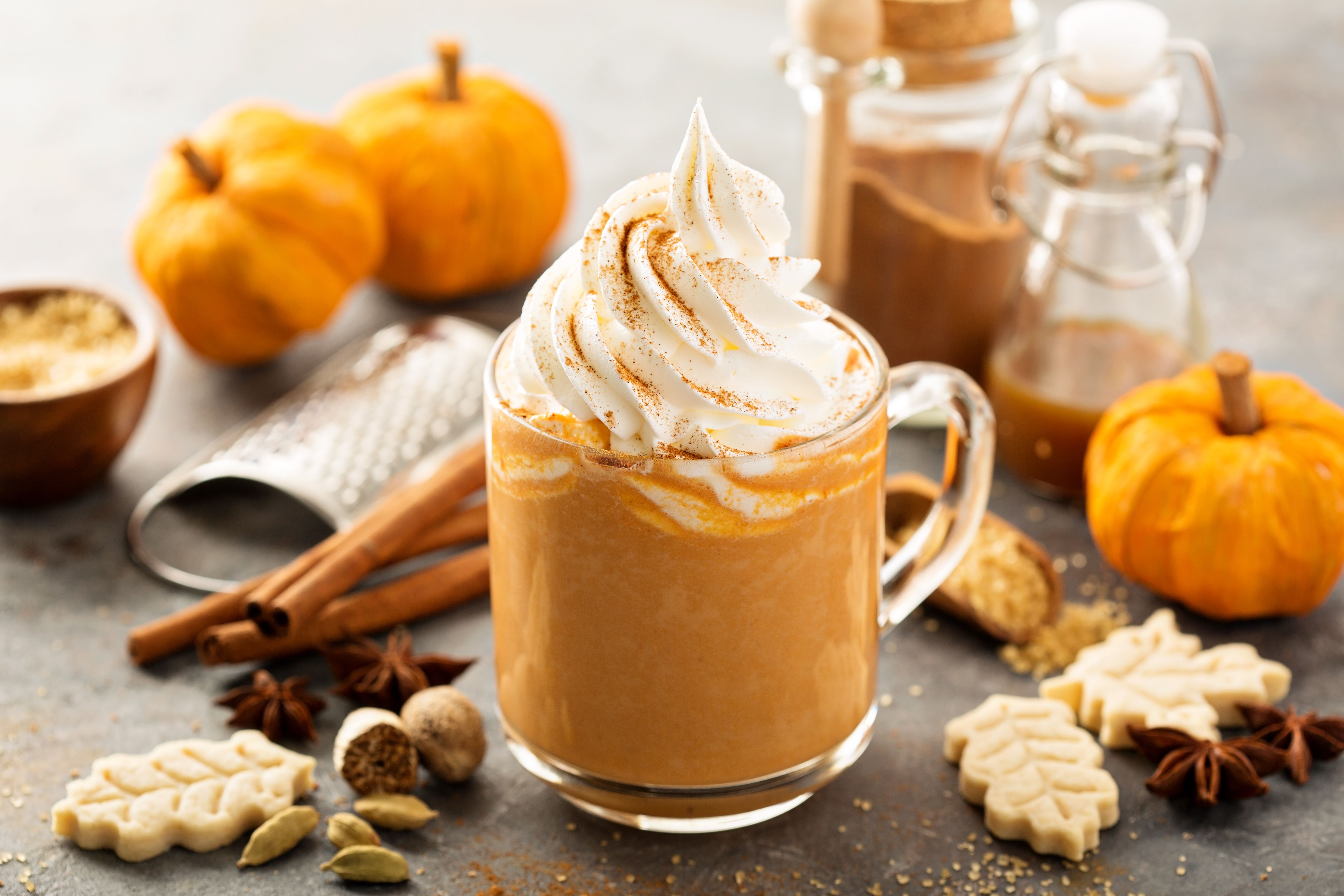 A pumpkin spice latte topped with whipped cream and cinnamon sits among autumn ingredients like pumpkins, cinnamon sticks, and fall-themed cookies. This image represents the seasonal appeal of pumpkin spice products, highlighting the opportunity for convenience stores to maximize fall sales by offering popular pumpkin spice and other seasonal flavors.